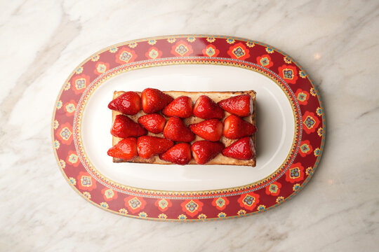 Close-up of a gourmet baked Alaska or meringue dessert topped with fresh juicy strawberries, served on an elegant red patterned platter over a marble table. Luxury restaurant sweet pastry