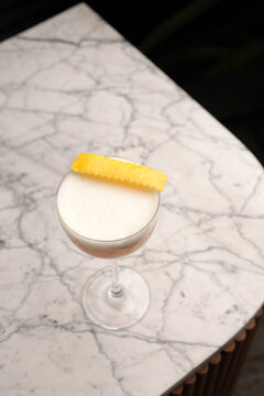 Classic sour cocktail with thick white foam and a yellow lemon twist garnish, served in a coupe glass on a marble table against a dark background. Fine dining bar drink