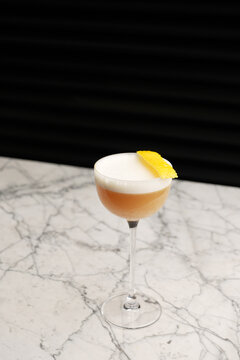 Classic sour cocktail with thick white foam and a yellow lemon twist garnish, served in a coupe glass on a marble table against a dark background. Fine dining bar drink