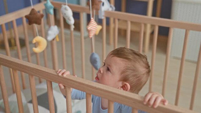 Boy crying with mouth open, gripping wooden crib rail and reaching toward hanging mobile in nursery; distress separation.