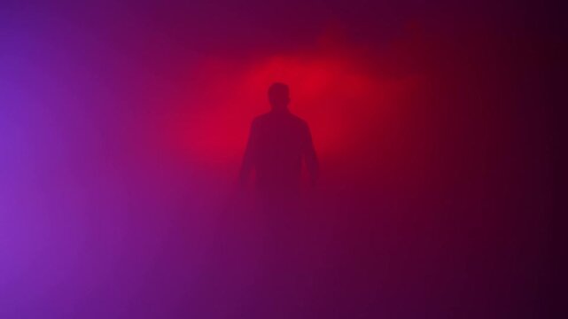 Slow dolly tracking shot of silhouetted man walking through dense fog in studio toward camera with red and purple backlight