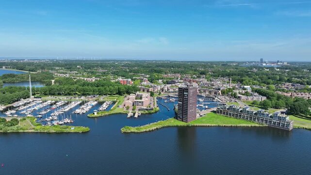 Aerial view of a marina with docked boats and a tall residential building surrounded by lush greenery and urban landscape in Almere, Flevoland, Netherlands.