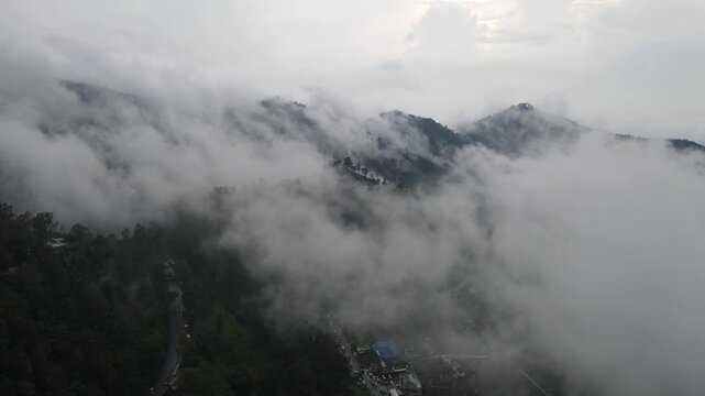 Aerial view of a winding mountain road passing through lush green hills and thick white clouds in Tawangmangu, Central Java, Indonesia.