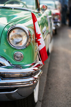 A classic car show is happening in downtown. People are walking around and looking at vintage vehicles. A green convertible is parked with Canadian flags and signs.