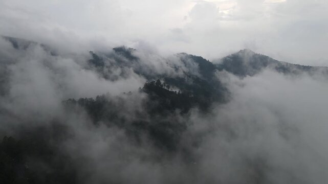 Aerial view of dense green forests and misty mountain peaks partially obscured by thick white clouds under an overcast sky in Tawangmangu, Central Java, Indonesia.