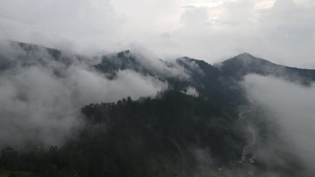 Aerial view of thick white mist rolling over dense green mountain forests under a cloudy sky in Tawangmangu, Central Java, Indonesia.