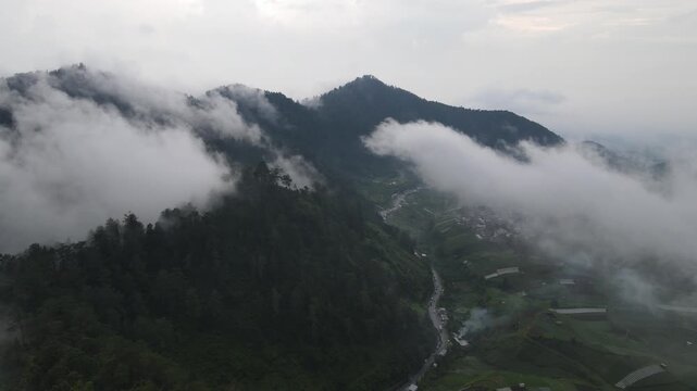 Aerial view of misty mountains and lush green hills with clouds rolling over the peaks and a winding road in the valley Tawangmangu, Central Java, Indonesia.