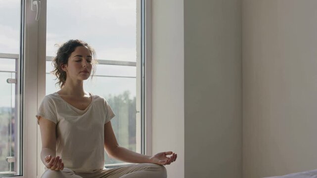 Young woman meditating in lotus pose with closed eyes on a sunny morning, practicing yoga and mindfulness for stress relief and mental health wellness near a window