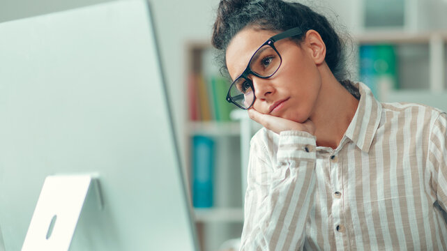Worried Young Woman With Glasses Staring At Computer Screen, Resting Head On Hand, Sitting At Desk In Modern Office. Focused On Work And Overwhelmed By Tasks, Business Concept