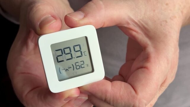 Close-up of woman's hands holding digital sensor displaying temperature and humidity, careful hands tracing display, wrinkled skin