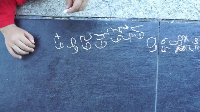 High-angle, top-down shot of a left-handed child in a red shirt sitting on the ground and writing a sequence of characters or numbers with white chalk on dark tiles.