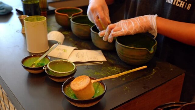 Gloved hands whisking matcha in ceramic bowl at tea counter. Close view of green tea powder, bamboo tools and dark pottery on workspace.