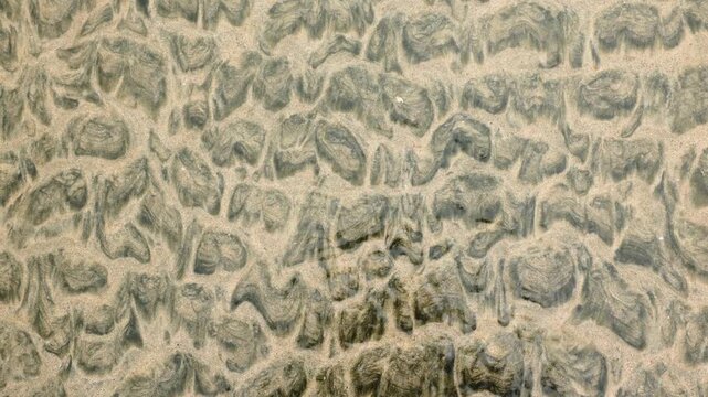 Close-up aerial view of wet beach sand revealing intricate natural ripple patterns and textures formed by receding tidal waters, creating mesmerizing organic wave-like formations on the shoreline.