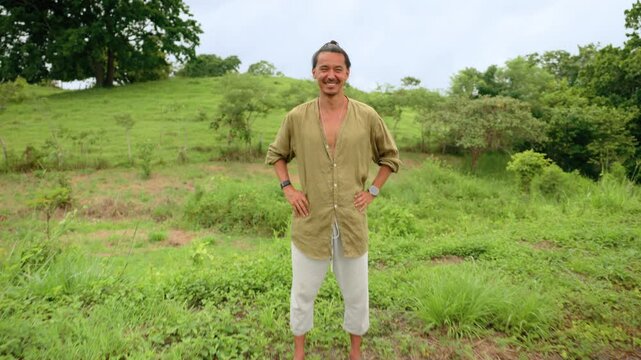 Happy man with hair bun wearing olive linen shirt smiles joyfully while standing in green rural meadow. Tropical hillside with trees and overcast sky creates natural backdrop.