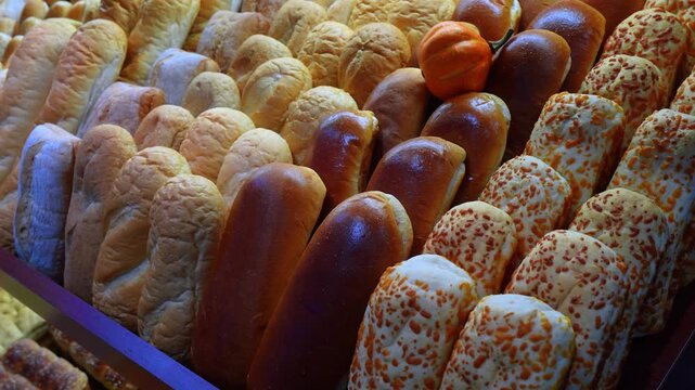 Close up view of assorted freshly baked bread rolls on a bakery shelf. A variety of golden buns, shiny brioche, and cheese topped loaves are arranged neatly with a small decorative pumpkin.