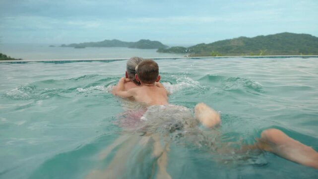 Father and son enjoy quality time swimming in turquoise infinity pool overlooking tropical mountains and ocean. Boy holds onto dad as they swim together in the clear water on vacation.
