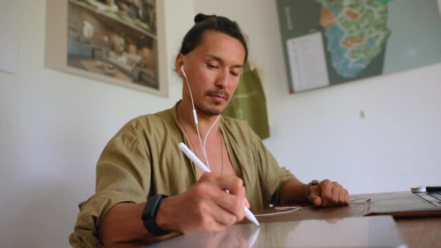 Creative professional with man bun works remotely at wooden desk, using stylus pen on tablet while listening through earphones, laptop nearby, maps and artwork decorating bright home office walls.
