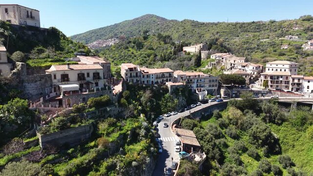 Aerial view of the historic hillside village of Savoca with stone buildings and a winding road nestled among lush green mountains in Savoca, Sicily, Italy.