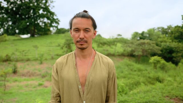 Serene man with top knot hairstyle wearing loose olive linen shirt and white pants stands peacefully in lush green tropical meadow surrounded by dense trees and rolling hills under cloudy sky.