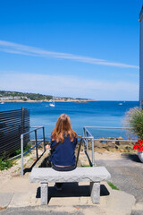 Fototapeta premium A woman sits on a stone bench overlooking a bright blue bay with sailboats, shoreline, stairs, railings, and flowers under a clear sky.
