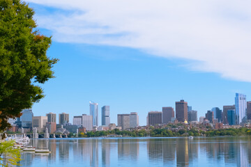 Fototapeta premium Boston skyline across the Charles river with calm water reflections, sailboats, trees, and a bright blue sky.