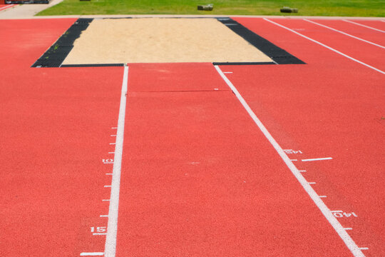 Red athletics runway with white distance markings leading toward a long jump sandpit bordered by black rubber and grass.