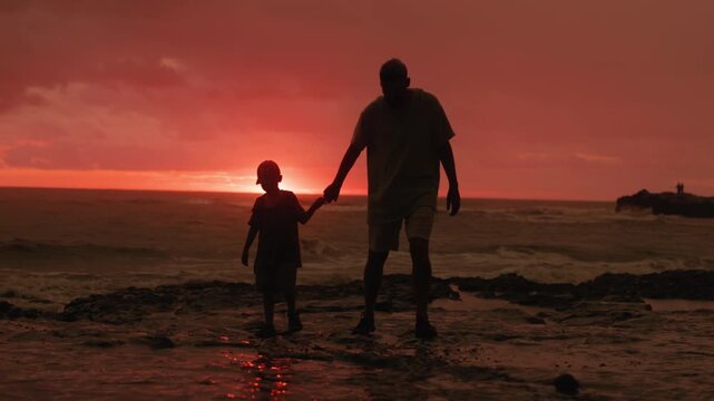 Father and son share a tender moment walking hand in hand across rocky tidal pools on the beach. Dramatic red and orange sunset sky creates stunning silhouettes against crashing ocean waves.