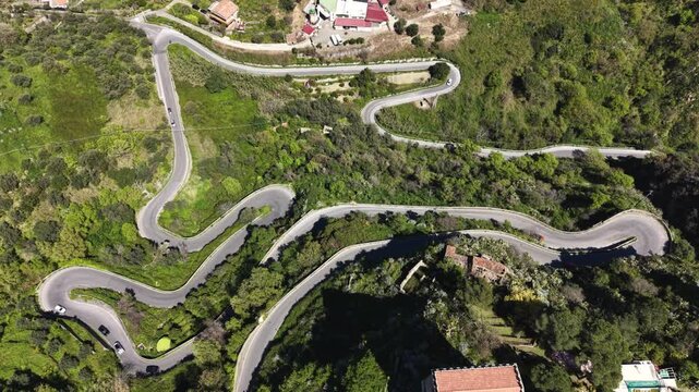 Aerial view of a winding mountain road with sharp hairpin turns through lush green hillsides and scattered buildings in Savoca, Sicily, Italy.