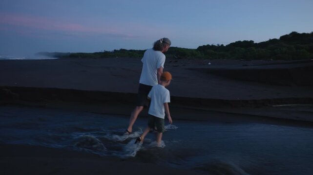 Father and young son wearing white shirts wade through gentle stream waters on dark volcanic black sand beach at dusk with pink and blue twilight sky, lush green hillside in background.