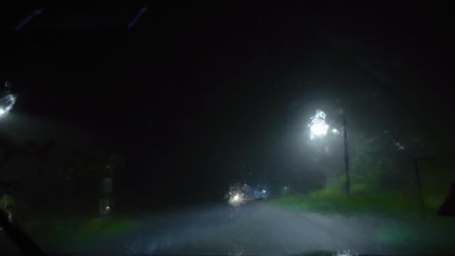Night road in heavy rain viewed through car windshield with headlights illuminating wet asphalt. Raindrops stream down glass as streetlights glow in darkness creating atmospheric driving conditions.