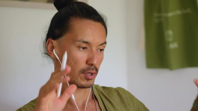 Business video call from home as man with top knot hairstyle speaks through white earphones while gesturing expressively. He wears olive green linen shirt and necklace in modern bright interior.