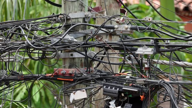 Tangled mess of black electrical cables and wires wrapped around weathered wooden utility pole with metal brackets and connectors, lush green tropical foliage visible in soft blurred background.
