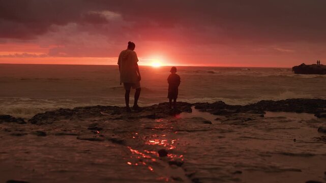 Father and son stand together on rocky shoreline watching dramatic sunset over ocean waves. Vivid orange and red sky reflects on tidal pools as silhouetted figures share peaceful moment by the sea.