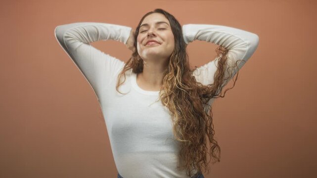 Woman with long wavy hair raising hands behind head against terracotta studio backdrop; serenity mindfulness.
