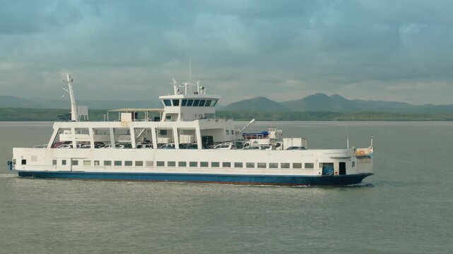 White and blue car ferry transports vehicles across calm ocean waters under cloudy skies. Tropical mountains and green coastline visible in background. Maritime transportation scene.
