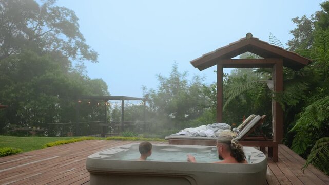 Father and son enjoy quality time together in steaming hot tub on wooden deck surrounded by lush tropical garden. Misty morning atmosphere with string lights visible on pergola in background.