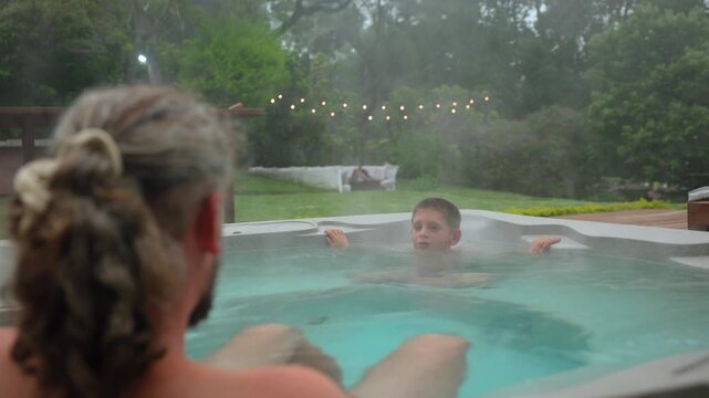 Father and son share quality time in a steaming hot tub on a backyard deck during evening hours. String lights glow softly as steam rises from warm turquoise water surrounded by lush green garden.