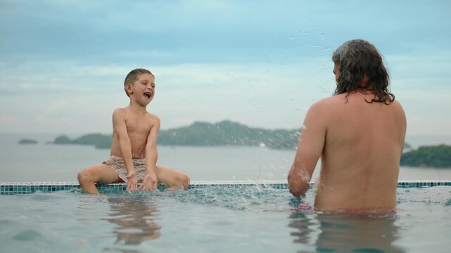 Joyful father and young son playing together in an infinity pool overlooking tropical islands. Boy sits on pool edge splashing water while dad watches from the turquoise water.