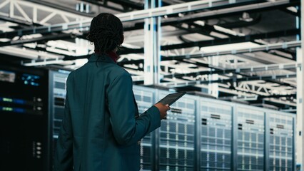 Obraz na płótnie Canvas Female programmer walking in data center using tablet to write code, compiling software patches, African American woman in server hub using device application to run programming script, camera B.
