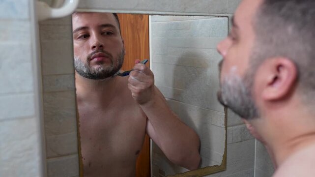 A young man shaving his beard in the bathroom in front of the mirror