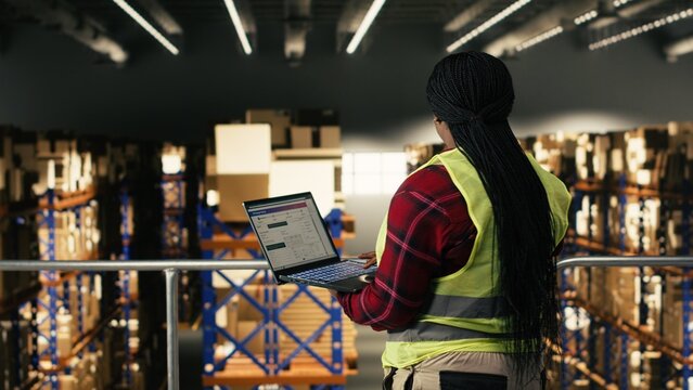 African American worker using laptop on elevated platform in fulfillment center, wearing high visibility safety vest, Employee manages logistics, tracking parcels and coordinating order fulfillment.