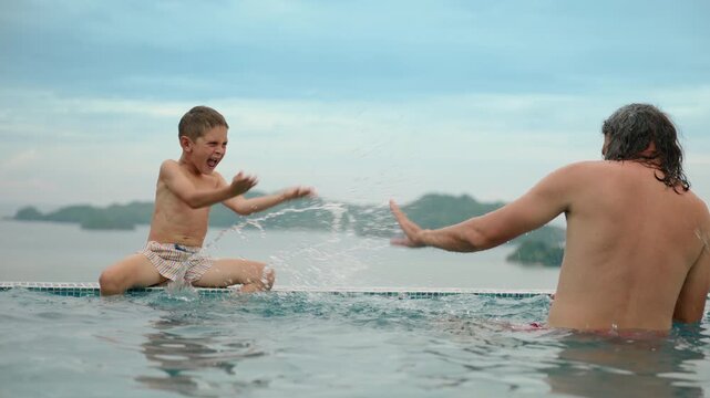 Happy father and son playing in infinity pool splashing water at each other. Boy sits on pool edge laughing while dad stands in water with tropical islands and mountains in background.