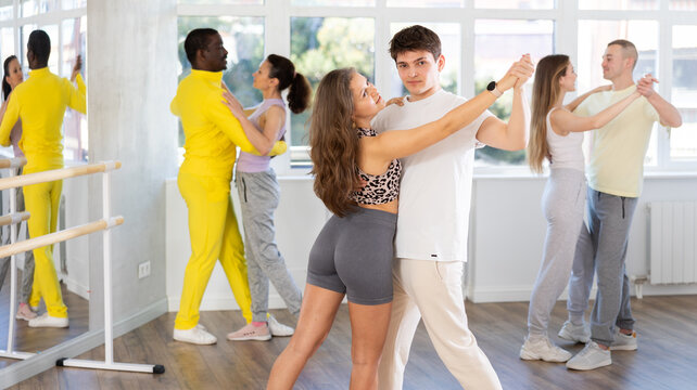 Smiling young, graceful girl and expressive Hispanic guy in sportswear enjoying slow foxtrot in dance studio. Amateur ballroom dancing concept