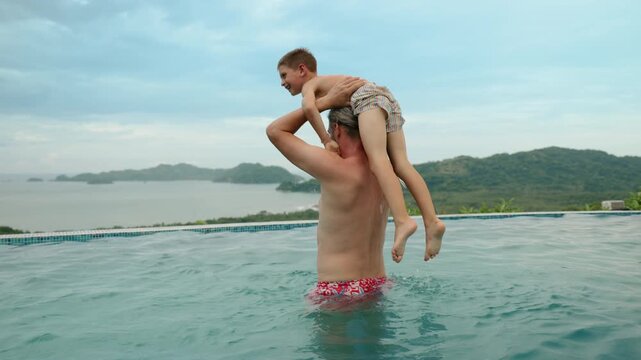 Father lifting and playing with his two children in a luxury infinity pool overlooking tropical mountains and ocean bay. Water splashes as kids laugh during family vacation fun.