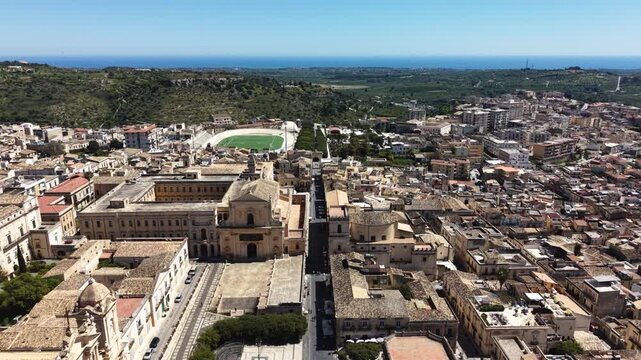 Aerial view of the historic Baroque architecture and rooftops of Noto with the cathedral and a stadium under a clear blue sky in Noto, Sicily, Italy.