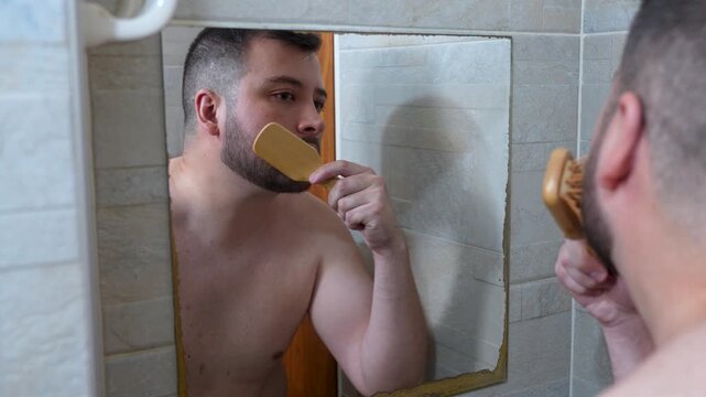 A young man combing his beard in the bathroom in front of the mirror