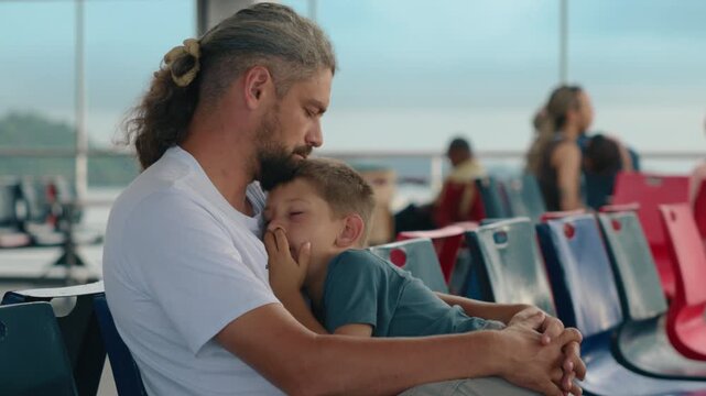 Father and son dozing peacefully on a ship as mountains pass by through windows. Bearded dad with long hair holds sleeping child in tender embrace on colorful ferry seats during family travel.