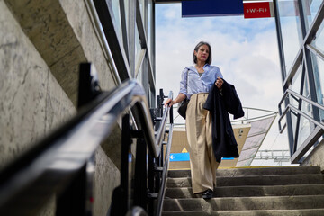 Businesswoman walking down stairs at a modern urban transit station, holding an overcoat and bag, representing daily commute, city life, and professional travel © DusanJelicic