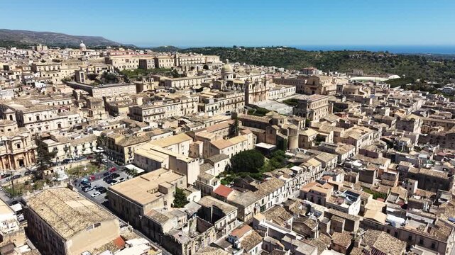 Aerial view of the historic Noto Cathedral surrounded by Baroque architecture and limestone rooftops under a clear blue sky in Noto, Sicily, Italy.