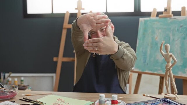 Man framing the view with hands, wearing apron beside an easel and canvas in a studio; creative concentration planning.